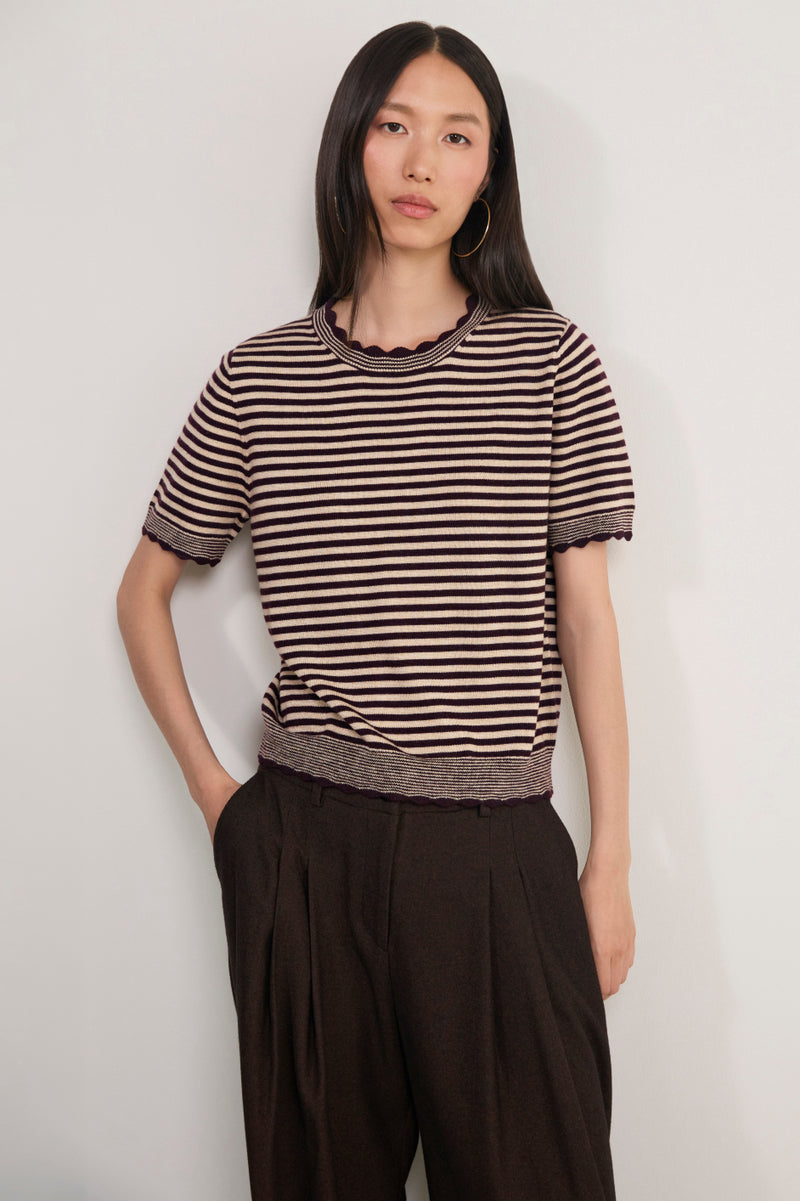 A woman modeling a short-sleeve knit top with horizontal brown and beige stripes, featuring a scalloped neckline and ribbed hem, paired with dark brown pleated trousers, against a plain light gray background.